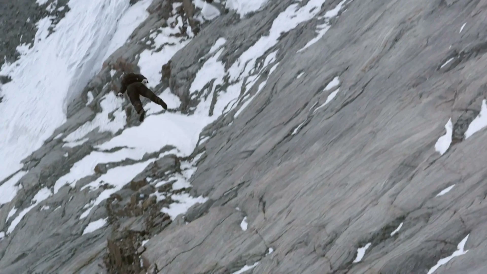 A person in a dark suit is seen falling down a steep, snow-dusted rocky mountain face, with a vast expanse of rock and snow.