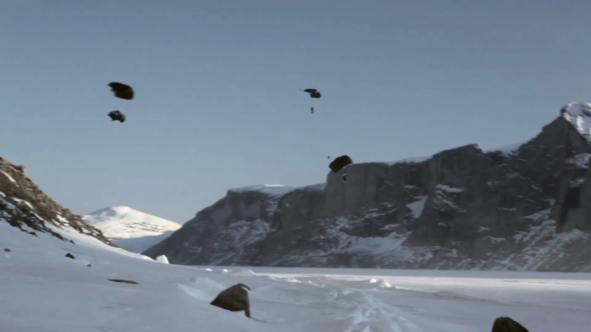 Four parachutists descend from a clear blue sky towards a vast, snow-covered landscape with towering mountains and a frozen lake.