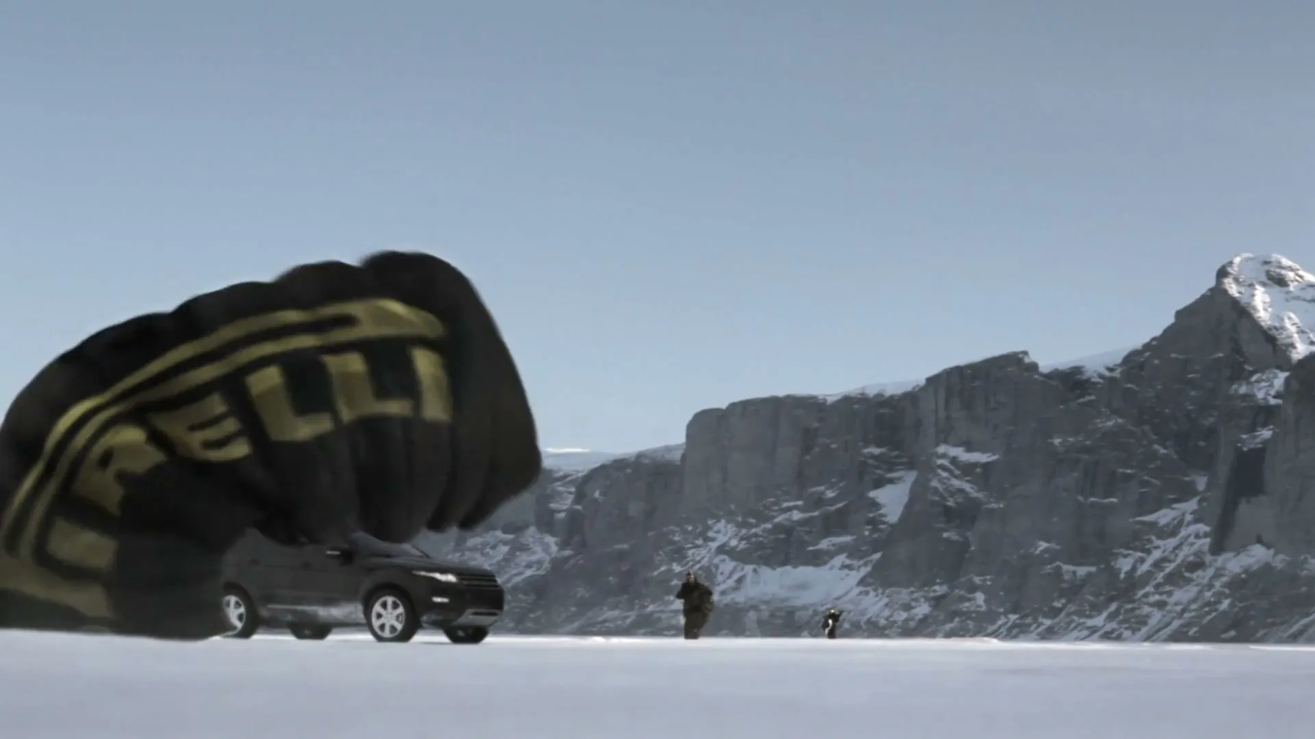 A black parachute with a gold Pirelli logo is dragged across a snowy plain by a black SUV, with two people running in the distance and mountains in the background.