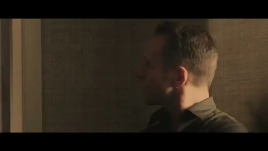 A man with short dark hair, wearing a dark shirt, looks to the left in a dimly lit room, appearing tense.