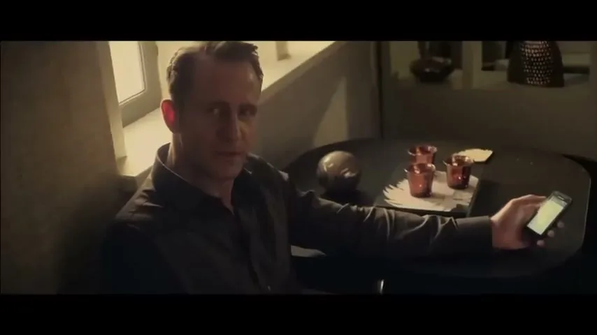 A man in his 40s, wearing a dark shirt, sits at a table, holding a smartphone and looking directly at the camera in a dimly lit interior.