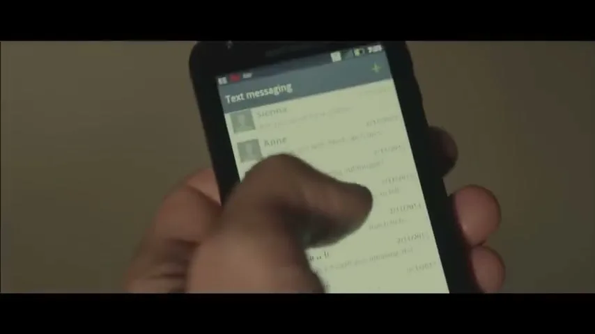 A person's hand scrolls through text messages on a smartphone, showing a list of contacts and dates on the screen.