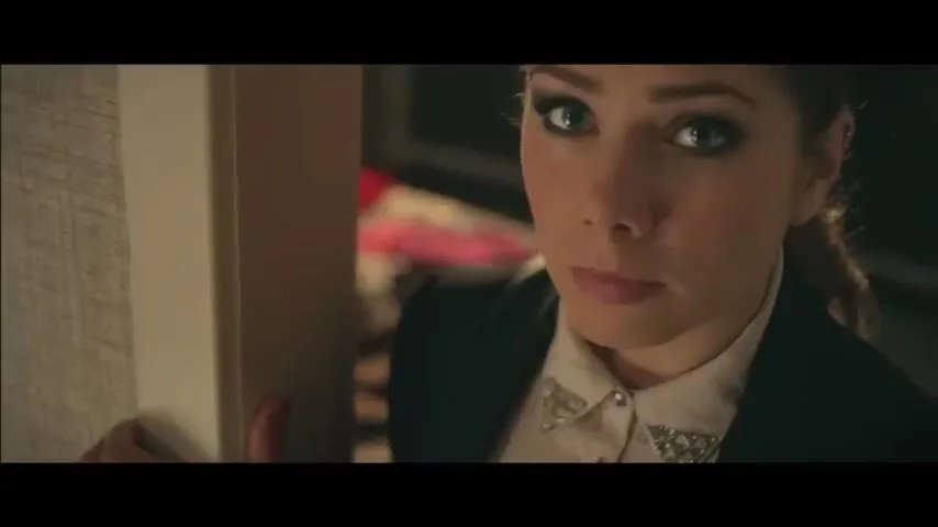 A young woman with dark hair and heavy eye makeup looks directly at the camera with a serious expression, partially obscured by a doorframe in an interior setting.