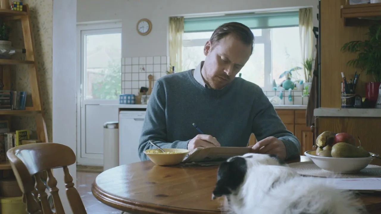 A man in a blue sweater sits at a kitchen table, writing on paper, while a black and white dog rests its head on the table next to him.