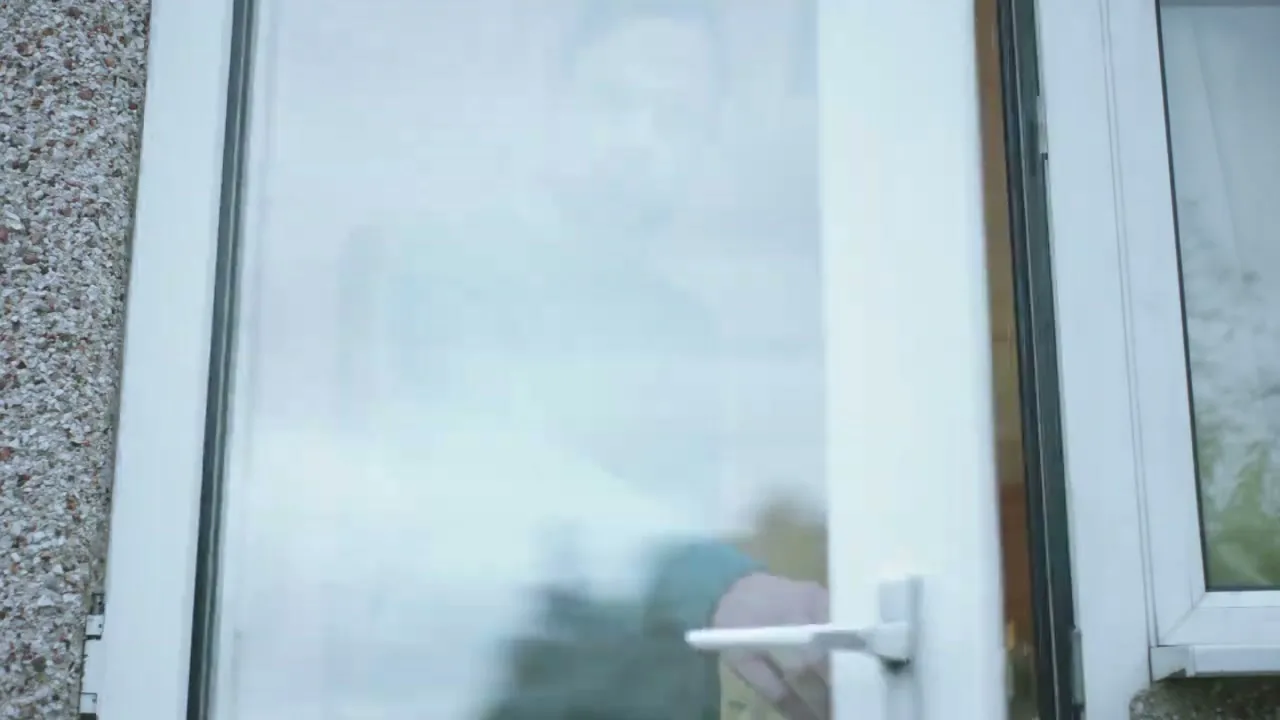 A man in a teal sweater opens a white door, looking out with a concerned expression from inside a house.