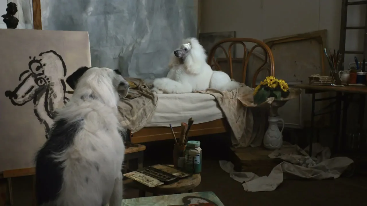 A black and white dog, an artist, paints a portrait of a white poodle posing on a bed in a messy artist's studio.
