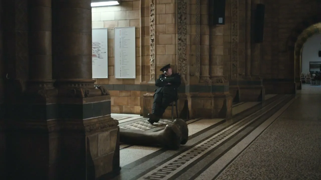 A male security guard in uniform is sleeping on a chair in a museum hallway next to a large dinosaur bone.