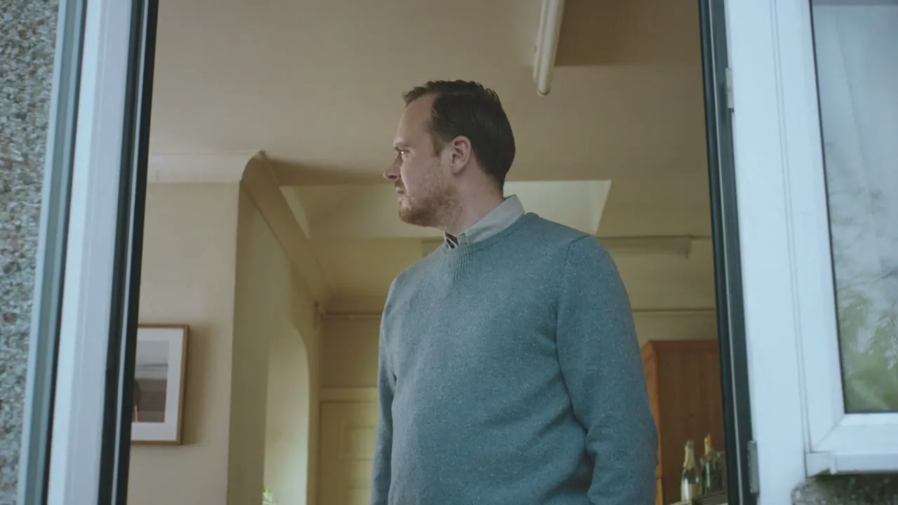 A man in a blue sweater stands in a doorway, looking around with a confused expression in a house during the day.