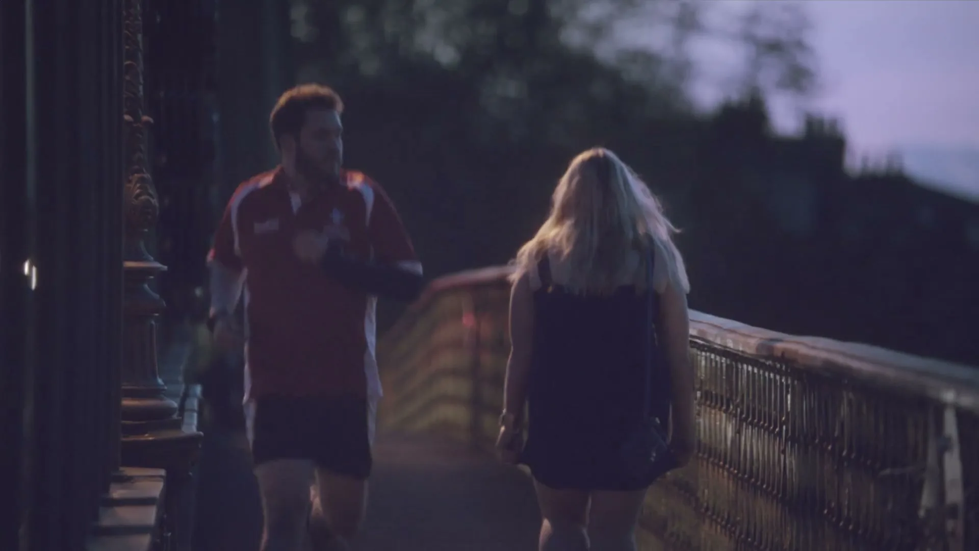 A blonde woman in a black dress walks away from the camera on a bridge at dusk, as a man in a red shirt jogs towards her.