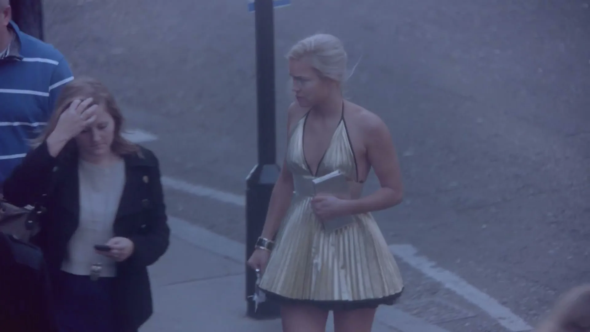 A young woman in a gold dress walks down a street, looking disheveled and carrying her shoes, as other people pass by.