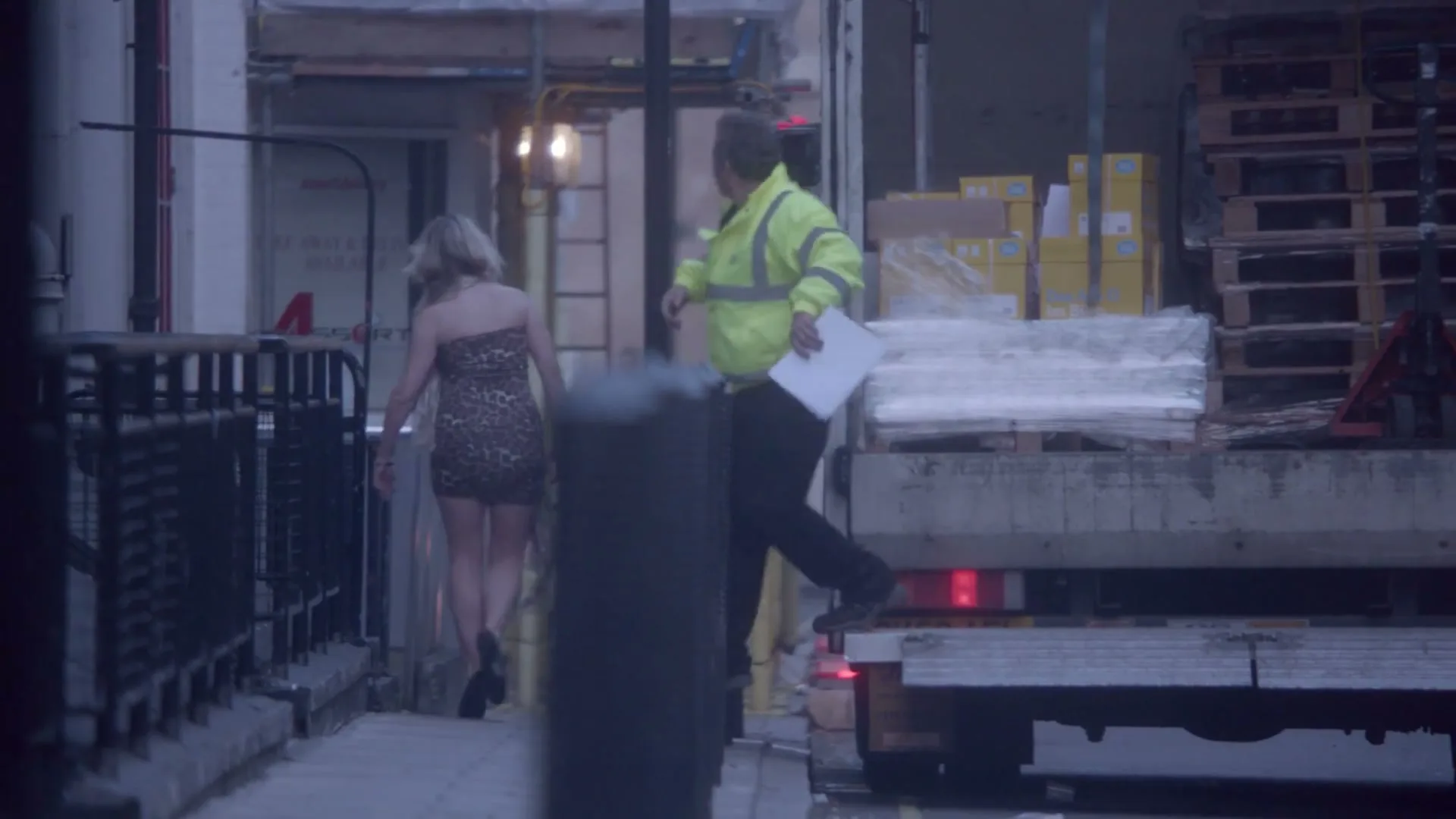 A young woman in a leopard print dress walks away from the camera on a city street, while a delivery man in a high-vis jacket stands on the back of a truck.