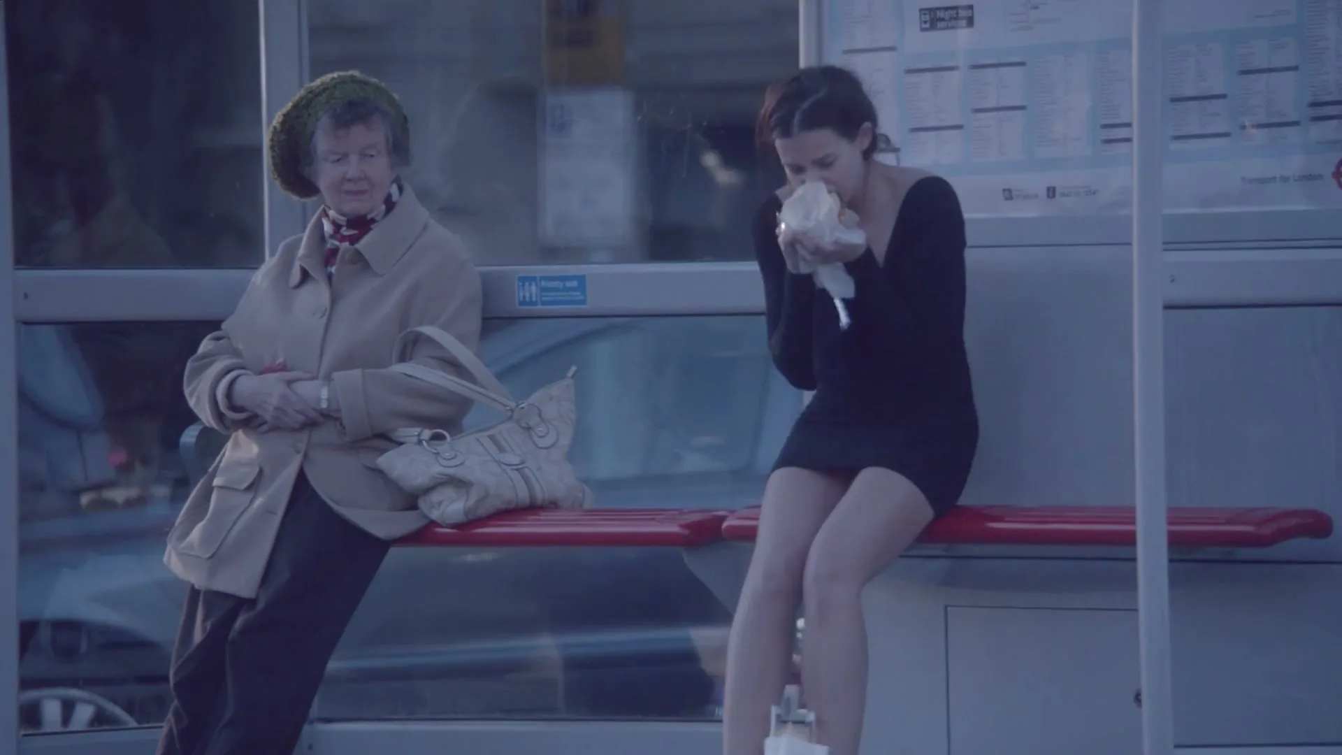 A young woman in a black dress sits on a bus stop bench, looking disheveled and holding a white bag to her face, next to an older woman.