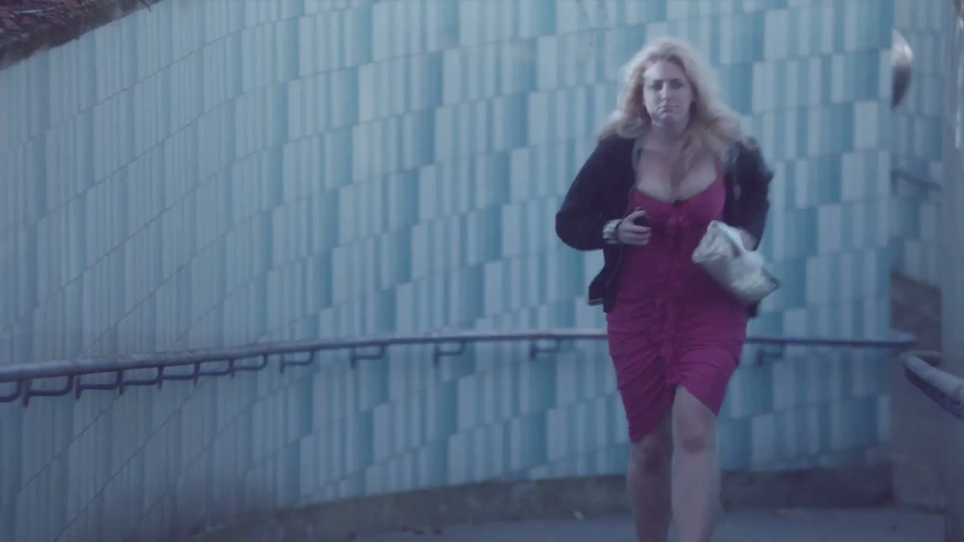 A disheveled blonde woman in a pink dress and black cardigan runs through a pedestrian underpass, clutching her chest and a white handbag.