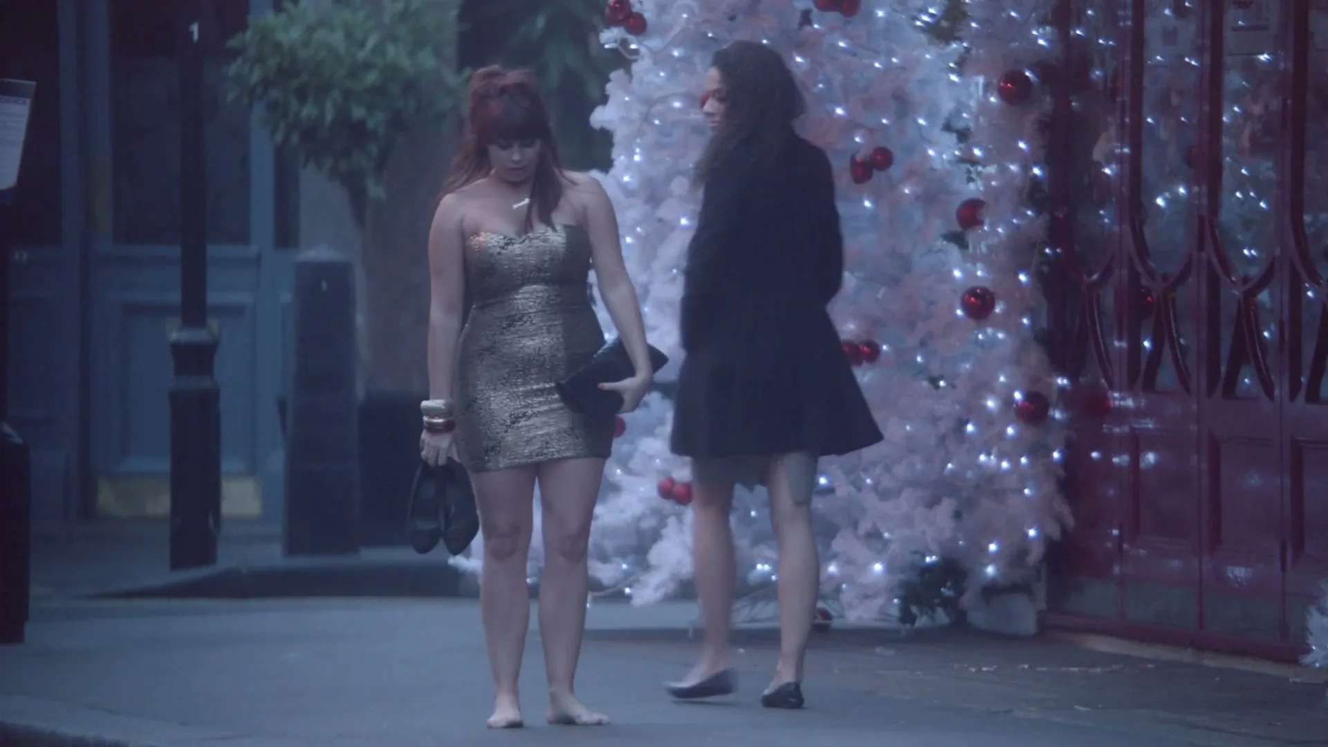 A young woman in a gold dress, holding her shoes and a clutch, walks barefoot on a street past a white Christmas tree at night.
