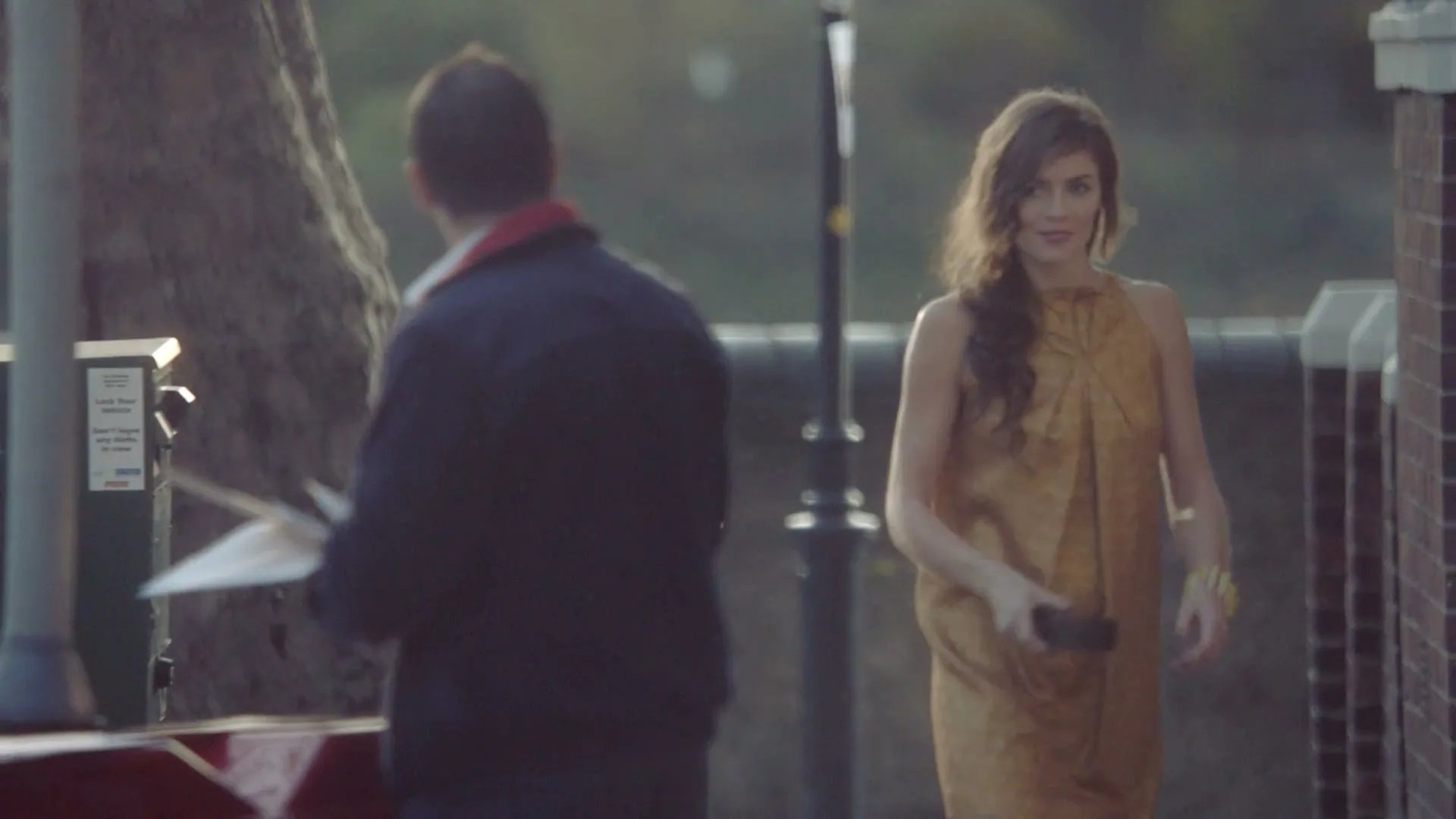 A young woman in a gold dress walks past a man on a sidewalk, then smiles as she approaches a gate in the golden hour.