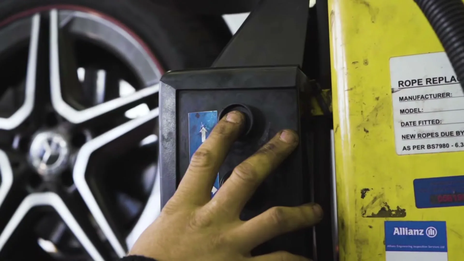 A mechanic's hand presses a button on a control panel, with a car wheel and yellow machinery visible in a garage.
