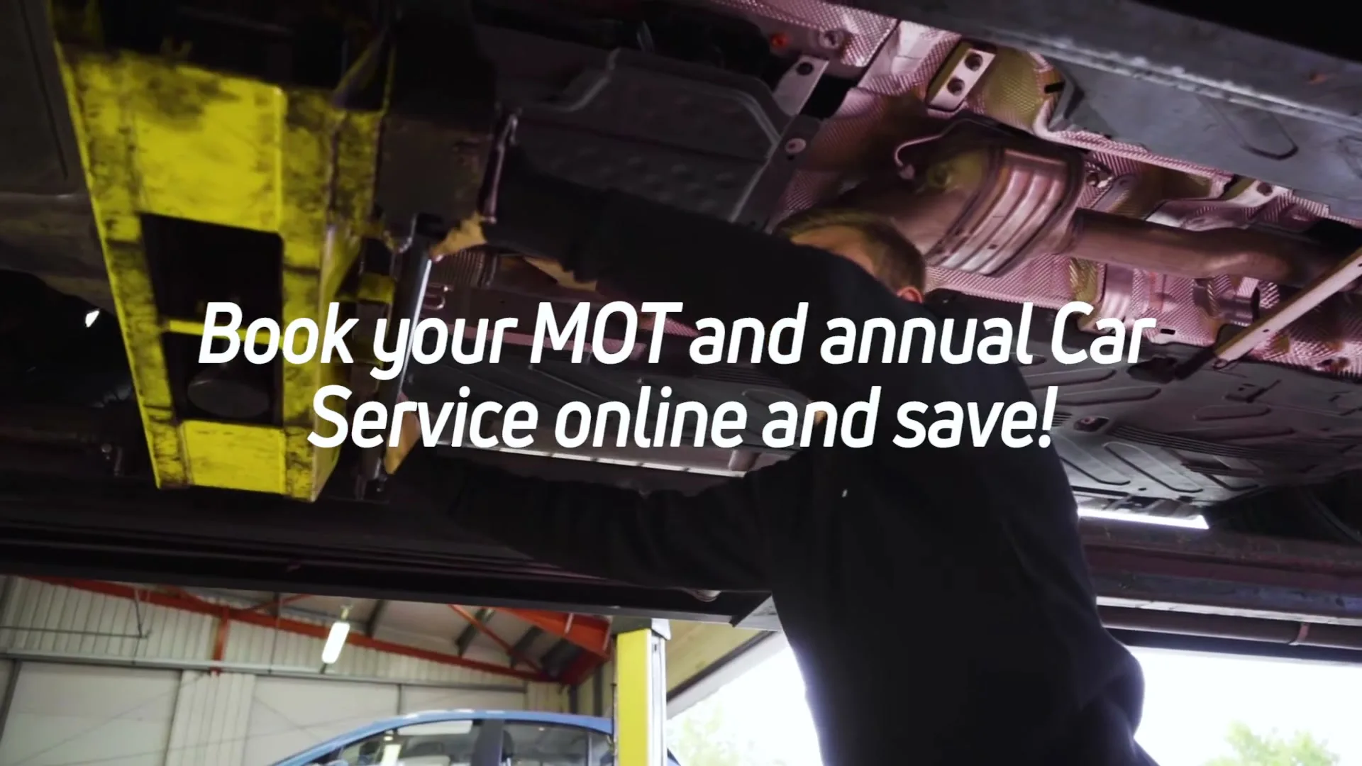 A male mechanic in a black uniform works on the underside of a car on a lift in a garage.