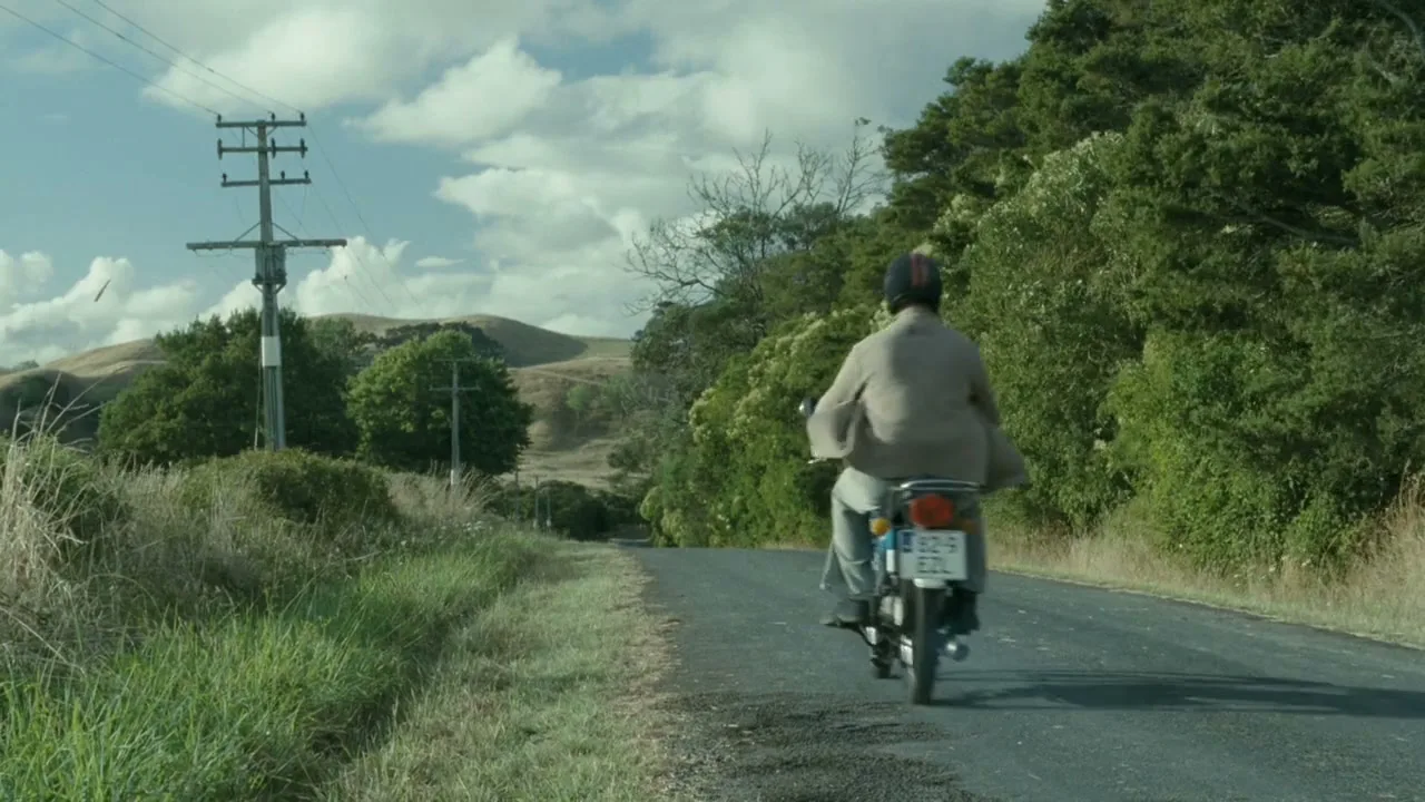A man in a helmet and jacket rides a small motorcycle away from the camera down a rural road lined with trees and power lines under a cloudy sky.