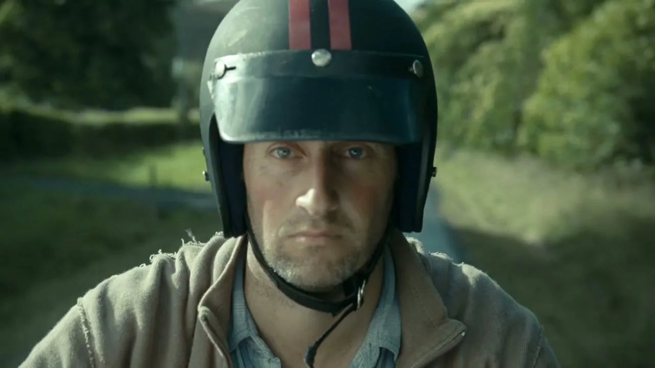 A man in his 30s, wearing a motorcycle helmet and a brown jacket, looks directly at the camera while riding a motorcycle on a rural road.