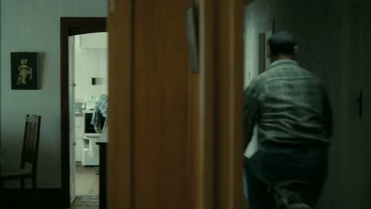 An older man in a plaid shirt and cap rides a small bicycle down a dimly lit hallway in a house.