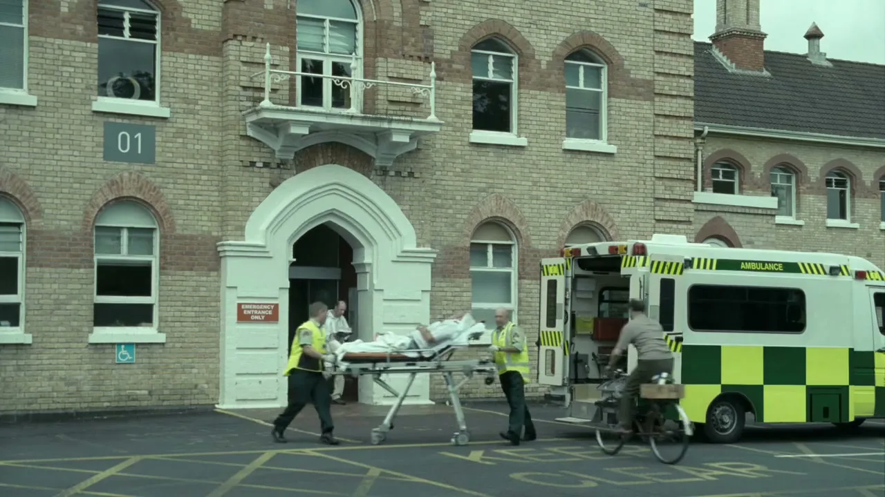 Two paramedics push a stretcher with a patient towards an ambulance parked outside a brick hospital building, while a man on a bicycle rides past.