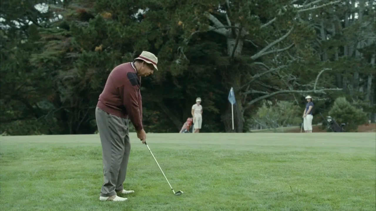 A man in a hat and sweater prepares to hit a golf ball on a green course, with other golfers in the background.