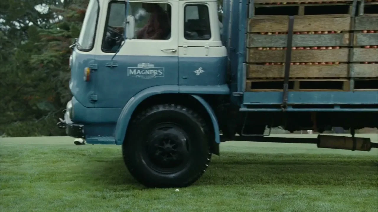 A vintage blue and white truck carrying crates of apples drives across a golf course, obscuring a golfer in a argyle sweater.