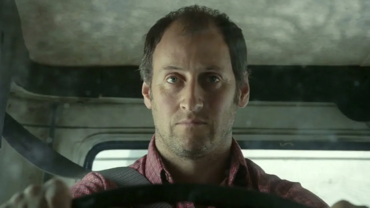 A man with short brown hair and a beard drives a vehicle, looking directly at the camera with a serious expression.