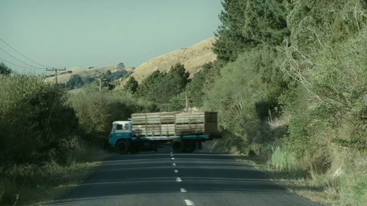 A blue and white truck carrying wooden crates drives past a silver car on a winding country road, surrounded by green bushes and hills under a clear sky.