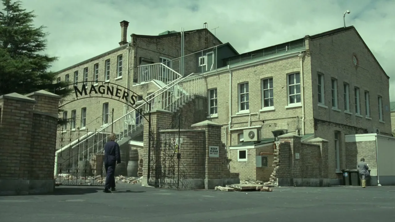 A wide shot of the Magners factory exterior, with two workers walking across the paved yard under a cloudy sky.