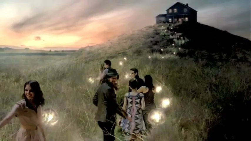 A diverse group of people, dressed in various outfits, walk along a glowing path through a grassy field towards a house on a hill at sunset.