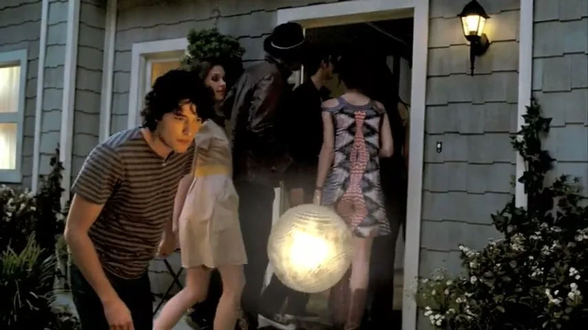 A group of young adults, dressed in casual attire, are entering a house at dusk, with a glowing orb floating nearby.