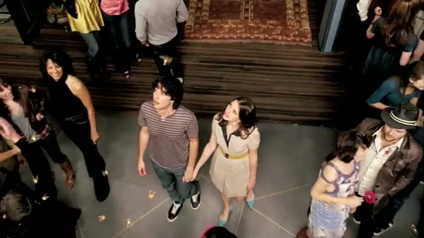 A young couple, holding hands, looks up in wonder as they stand amidst a diverse crowd of people dancing and socializing at a lively party.