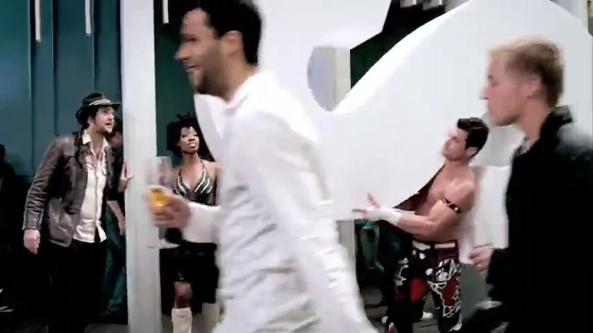 A diverse group of people, including a man in a fedora and a woman in a black dress, stand in a room with large white letters, as a man in a white shirt walks by.