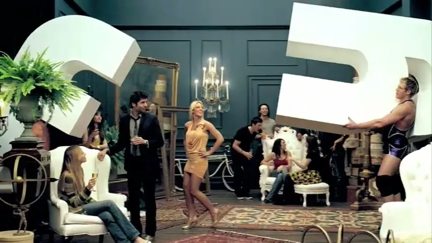 A diverse group of people are at a party in a lavish room, with a man in a wrestling singlet carrying a giant letter 'E' and a woman in a gold dress dancing.