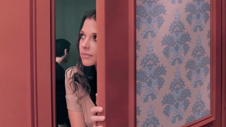 A young woman with long dark hair peeks out from behind a red door with patterned wallpaper, looking cautiously to her left.