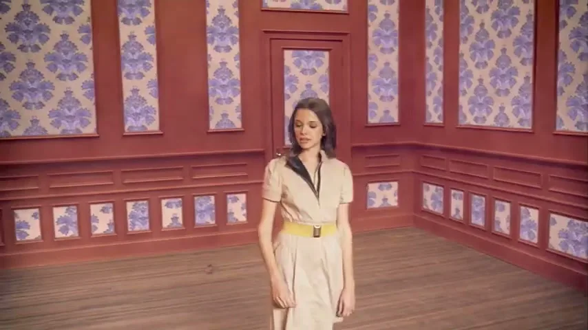 A young woman in a beige dress with a yellow belt stands in a room with red wainscoting and purple patterned wallpaper, looking around curiously.