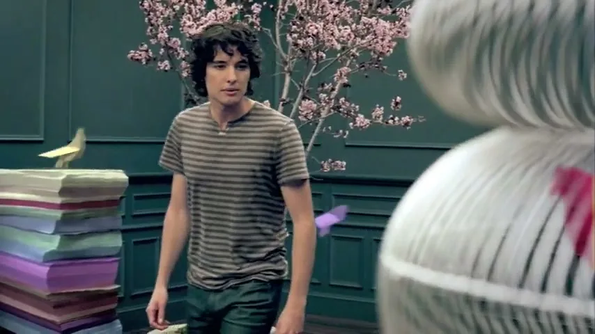 A young man with curly hair, wearing a striped shirt, looks curiously at a large, white, abstract birdcage in a room with a cherry blossom tree and a stack of colorful books.