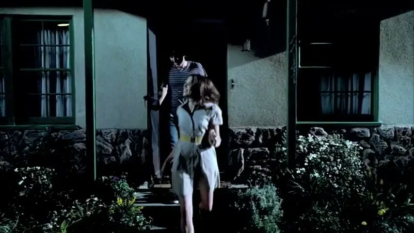 A young woman in a dress and a young man in a striped shirt run out of a house at night, looking scared.