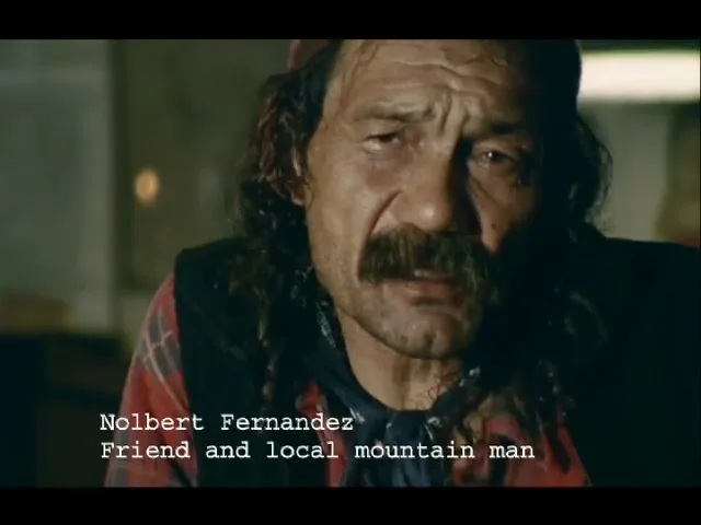 A man with a mustache and long hair, wearing a red plaid shirt and a beanie, looks directly at the camera in an interior setting.