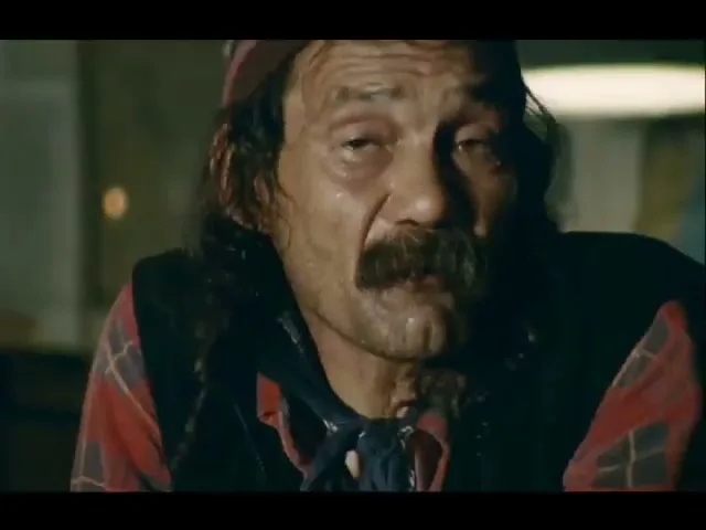 A man with a mustache and long hair, wearing a plaid shirt and a patterned hat, looks tired and sweaty in an interior setting.