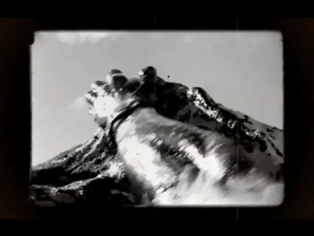 A person's hands, covered in mud, are shown gripping a rocky surface as they climb a mountain under a bright sky.