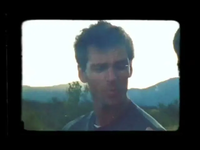 A man with dark hair and a grey shirt smiles and talks, then the shot quickly cuts to a blurry, scratched view of the sky and grass.