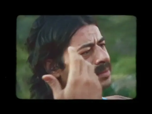 A man with dark hair and a mustache looks off-camera with a concerned expression, touching his temple, in an outdoor setting.