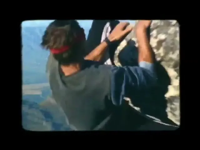 A blindfolded man in a t-shirt and jeans climbs a rocky mountain face, with a vast landscape visible in the background.