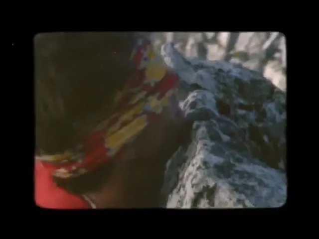 A person with a colorful bandana and red shirt is seen from the side, climbing a rocky mountain face on a sunny day.