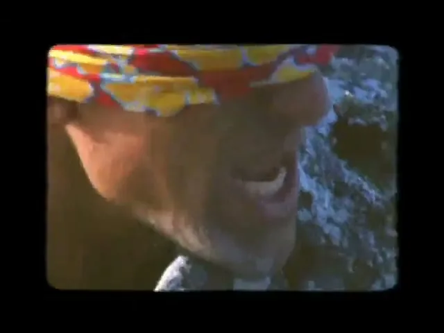 A man with a colorful bandana and long hair is seen in a medium closeup, grimacing while climbing a rocky mountain.