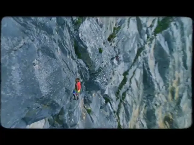 A male rock climber in a red top and colorful shorts scales a steep, grey rock face under a bright sky.