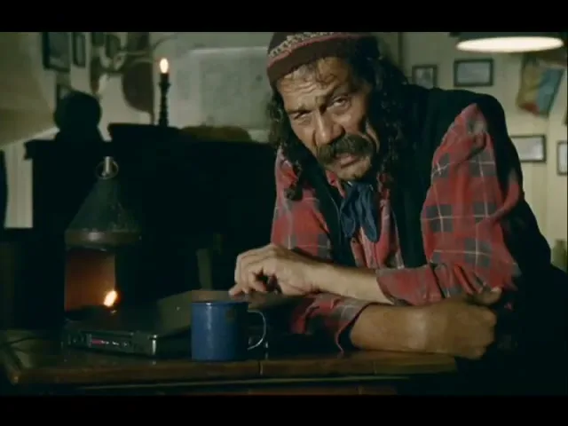 A man with a mustache and long hair, wearing a red plaid shirt and a beanie, sits at a wooden table and opens a laptop in a dimly lit room.