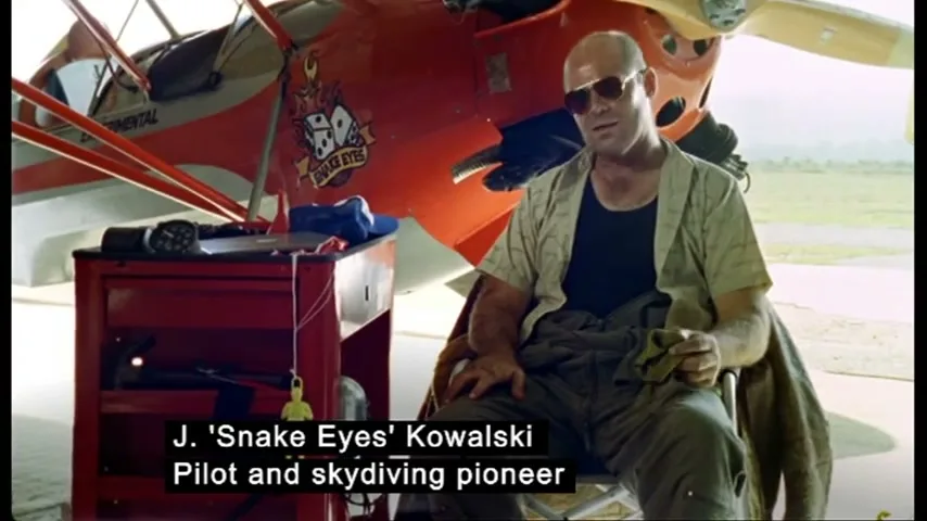 A bald man in sunglasses and a casual shirt sits in a chair next to a red biplane, holding a cloth, in an airplane hangar.